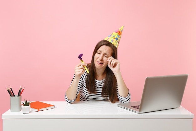 Mulher chorando com chapéu de festa de aniversário tocando cachimbo comemorando sozinha enxugando as lágrimas enquanto está sentada trabalhando na mesa branca com o laptop do pc