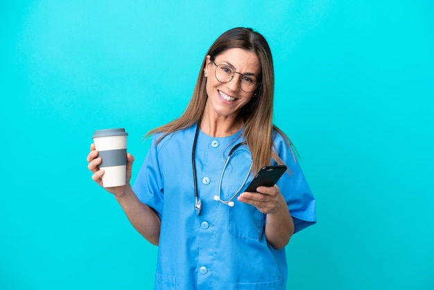 Mulher cirurgiã de meia idade isolada em fundo azul segurando café para levar e um celular