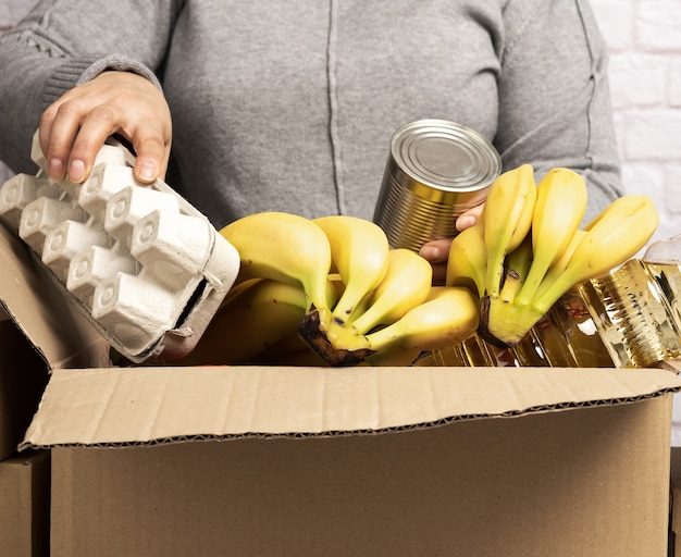 Mulher coleta alimentos, frutas e coisas em uma caixa de papelão para ajudar quem precisa, ajuda e conceito de voluntariado. entrega de produtos