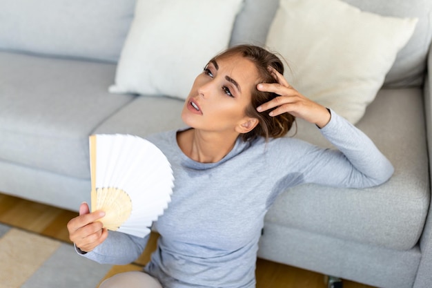 Mulher coloca a cabeça nas almofadas do sofá se sente lenta devido às ondas de calor insuportáveis ventilador de mão se refrescar apartamento quente de verão sem ar condicionado conceito de sistema de controle climático