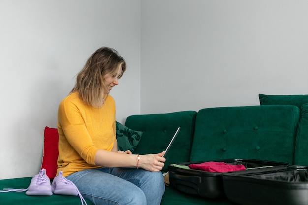 Mulher coloca roupas em uma mala preta sentada no sofá na sala de casa segurando um tablet mulher feliz dobra roupas