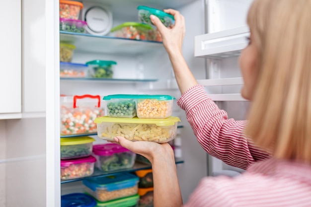 Mulher colocando recipientes com legumes misturados congelados do freezer.