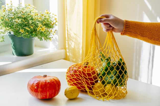 Mulher com a mão segurando uma rede de compras com legumes, abóboras e salada verde na cozinha Cores amarelas e laranjas Conceito de desperdício zero