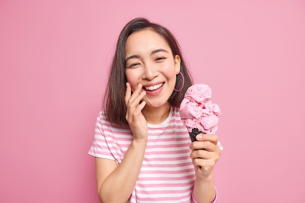 mulher com aparência oriental sorri com os dentes vestida com uma camiseta listrada casual segurando sorvete de waffle e está de bom humor