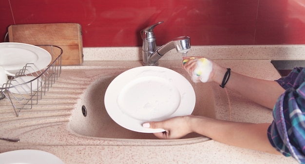 Mulher com as mãos lavando um prato branco na pia da cozinha.