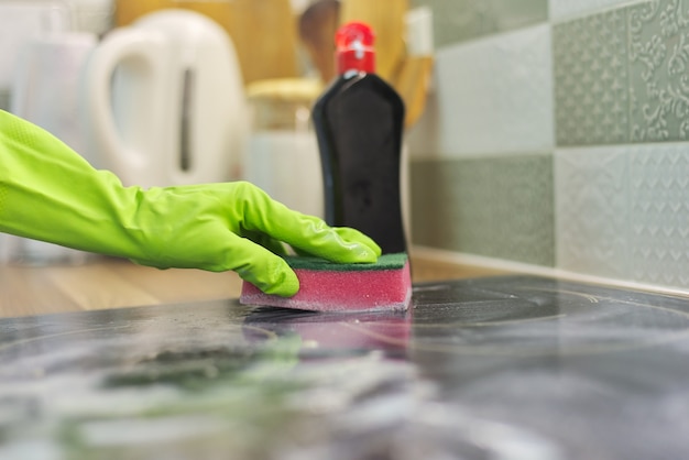 Mulher com as mãos nas luvas limpa a placa de vitrocerâmica da cozinha com esponja e detergente