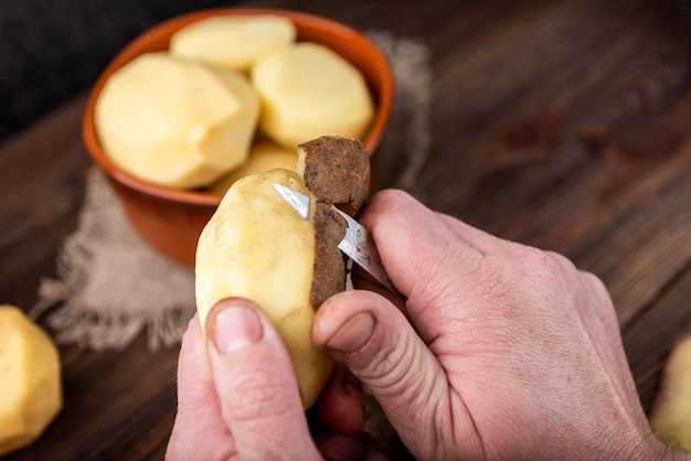 Mulher com as mãos para descascar batata. Limpe as batatas na superfície de madeira. Reparar alimentos para cozinhar.