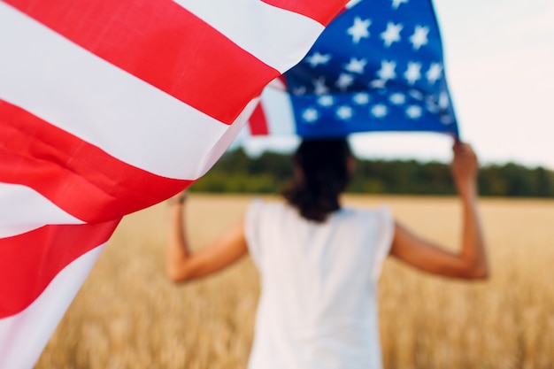 Mulher com bandeira americana no campo de trigo ao pôr do sol. 4 de julho. Feriado patriótico do Dia da Independência.
