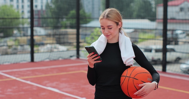Mulher com bola de basquete e smartphone na quadra de basquete Usando smartphone após o treinamento Conceito de estilo de vida ativo do esporte
