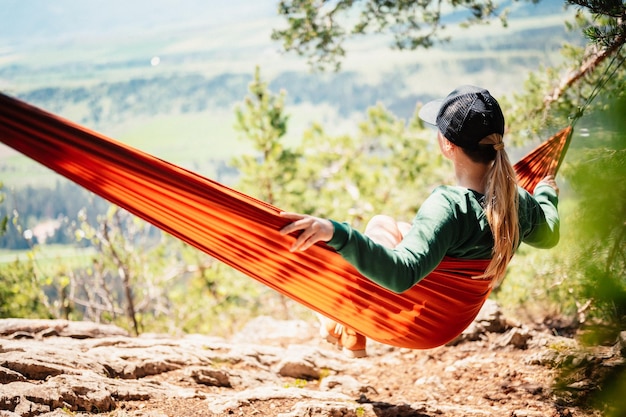 Mulher com boné descansando em confortável rede nas montanhas relaxando na rede laranja entre duas árvores de pinho apreciando a vista da natureza