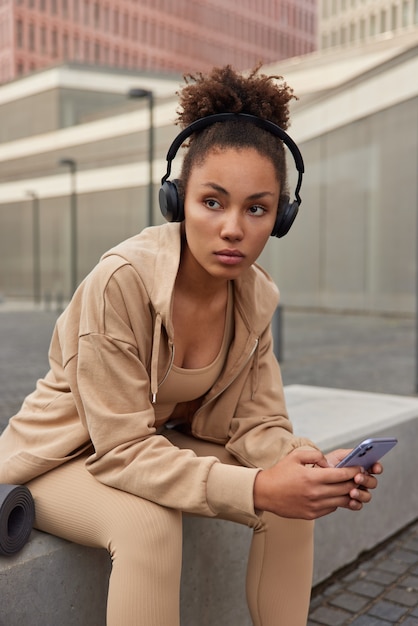 mulher com cabelo afro faz uma pausa após o treino baixa músicas para a lista de reprodução ouve música desviando o olhar poses em ambiente urbano