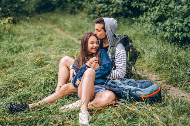 Mulher com cabelo comprido e homem sentado na grama na floresta com mochilas. Divirta-se na natureza, abraçando e beijando