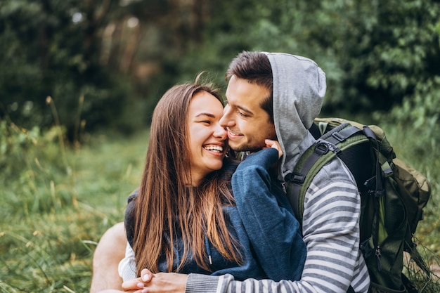 Mulher com cabelo comprido e homem sentado na grama na floresta com mochilas. Divirta-se na natureza, abraçando e beijando