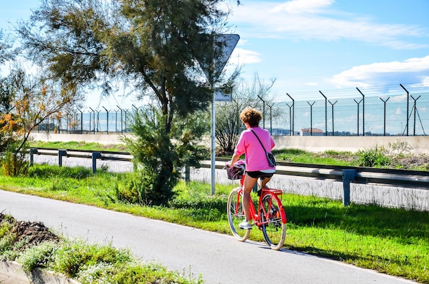 Mulher com cabelo curto e encaracolado, andando de bicicleta vermelha com uma cesta, em uma ciclovia com árvores.