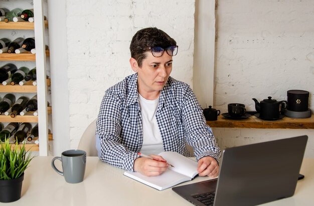 Mulher com cabelo curto se senta à mesa no escritório, trabalhando no computador, olhando para a tela com surpresa, levantando os óculos na testa. Mulher fazendo anotações em um caderno, trabalhando em um escritório doméstico com um laptop
