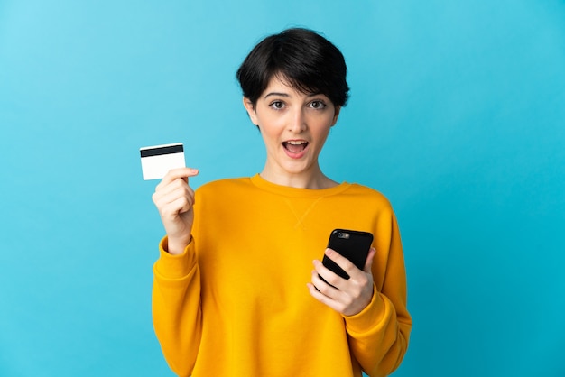 Mulher com cabelo curto sobre parede isolada comprando com o celular e segurando um cartão de crédito com expressão de surpresa