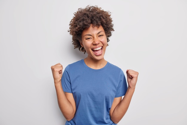 mulher com cabelo encaracolado aperta os punhos alegra-se sucesso alcança prêmio vestida com camiseta azul casual isolada no branco. Modelo feminina sortuda torna-se campeã