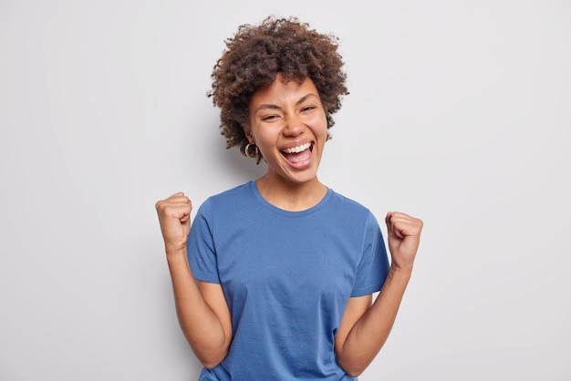 Mulher com cabelo encaracolado aperta os punhos alegra-se sucesso alcança prêmio vestida com camiseta azul casual isolada no branco. modelo feminina sortuda torna-se campeã