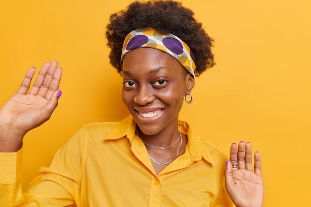 mulher com cabelo encaracolado dançando mantém as palmas das mãos levantadas e sorrisos amplamente usa camiseta casual com bandana amarela