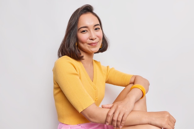 mulher com cabelo escuro parece satisfeita com a câmera sentada relaxada usa macacão amarelo e pulseira fica depois de caminhar escuta interlocutor com prazer isolado no branco