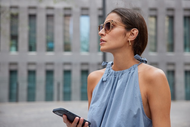 mulher com cabelo escuro usa telefone celular para bater papo online usa óculos escuros da moda e blusa azul posa contra um prédio na rua