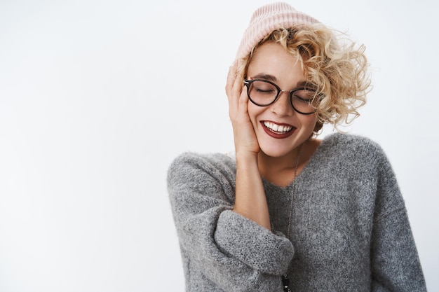 mulher com cabelo loiro curto encaracolado no gorro, óculos, blusa aconchegante, sorrindo com amor e deleite, tocando a bochecha, feche os olhos.