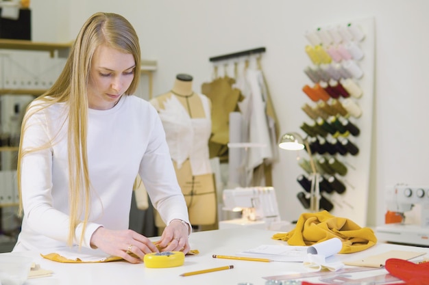 Mulher com cabelo loiro freelancer designer de moda ou alfaiate trabalhando em um projeto ou rascunho com tecidos coloridos na oficina vista frontal