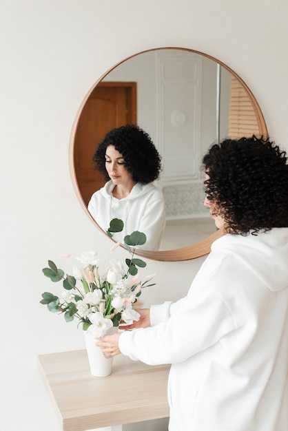 Mulher com cabelo preto encaracolado coloca um buquê de flores em um vaso branco na mesa