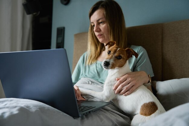 Mulher com cachorro fofo deitado na cama e usando laptop de manhã