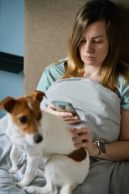 Mulher com cachorro relaxando na cama usa smartphone