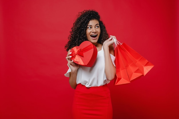 Mulher com caixa de presente em forma de coração e sacos de compras na parede vermelha