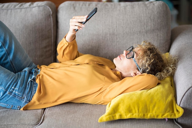 Mulher com camisa amarela aproveita o tempo e o lazer em casa deitada no sofá e usando a conexão do celular Pessoas e tecnologia A senhora moderna se comunica e usa conta de mídia social e página da web