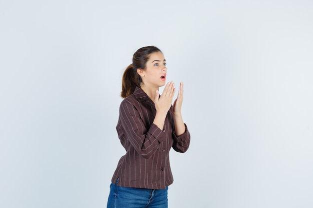 mulher com camisa listrada marrom com as mãos perto da boca aberta e parecendo chocada, vista frontal.