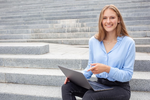 Mulher com cara de feliz sentada na escada e apontando para a tela do laptop olhando para a câmera