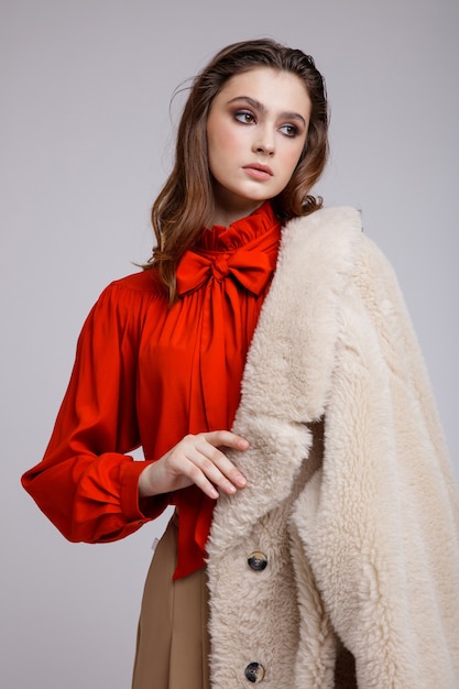 Mulher com casaco bege, saia longa, botas de blusa com laço vermelho no fundo branco. Cabelo castanho Fotografia de Estúdio