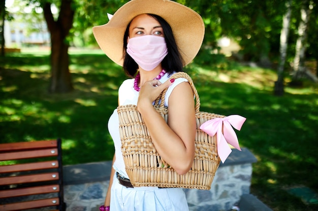 Mulher com chapéu de palha e bolsa usando máscara protetora rosa, no parque ao ar livre na cidade, autocuidado de conceito, vida durante a pandemia de coronavírus, covid-19.