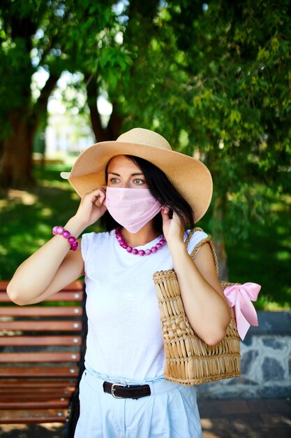 Mulher com chapéu de palha e bolsa usando máscara protetora rosa, no parque ao ar livre na cidade, autocuidado de conceito, vida durante a pandemia de coronavírus, covid-19.