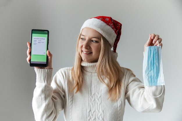 Mulher com chapéu de Papai Noel segurando telefone com certificado de vacinação cobiçoso e máscara médica protetora