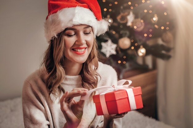 mulher com chapéu de Papai Noel sorrindo e segurando uma caixa de presente vermelha