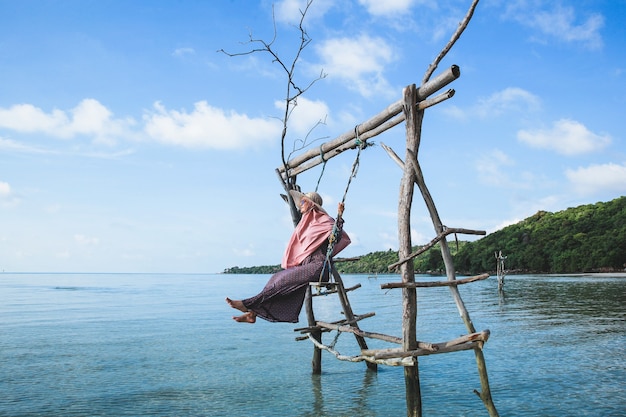 Mulher com chapéu de verão balançando em um balanço de madeira sobre a água do mar rasa e calma na ilha Karimun Jawa com um lindo fundo de céu azul