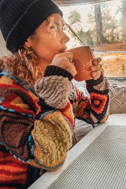 Foto mulher com chapéu e suéter pacwork sentada em uma caravana bebendo chá quente olhando pela janela conceito de relaxamento e lazer