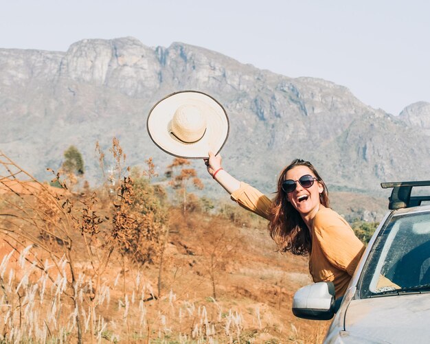 Foto mulher com chapéu na mão com corpo saindo do carro com serra do caraca ao fundo