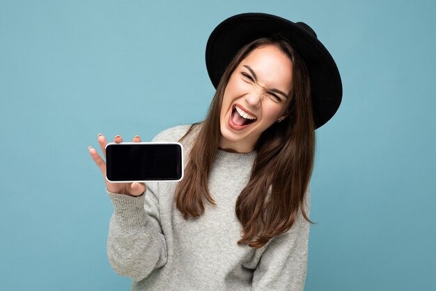 mulher com chapéu preto e suéter cinza se divertindo segurando o telefone, mostrando o smartphone isolado no fundo