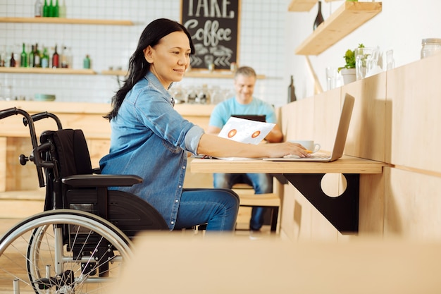 Mulher com deficiência e cabelos escuros, muito sorridente, sentada em uma cadeira de rodas, segurando uma folha de papel e trabalhando em seu laptop em um café