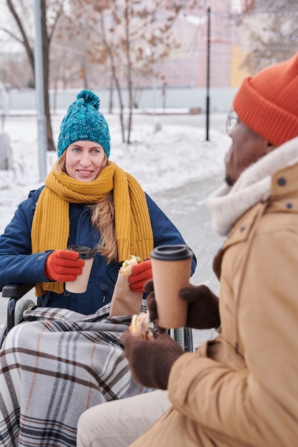 Mulher com deficiência em cadeira de rodas tomando café quente com um homem africano no parque em um dia de inverno