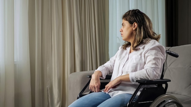 Mulher com deficiência triste em cadeira de rodas olha pela janela em casa