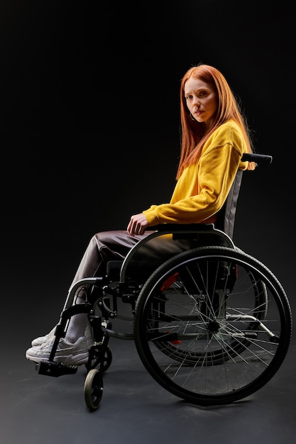 Mulher com deficiência triste em uma cadeira de rodas, olhando para a câmera deprimida. mulher ruiva na camisa amarela casual senta-se isolada no fundo preto. conceito de saúde e pessoas