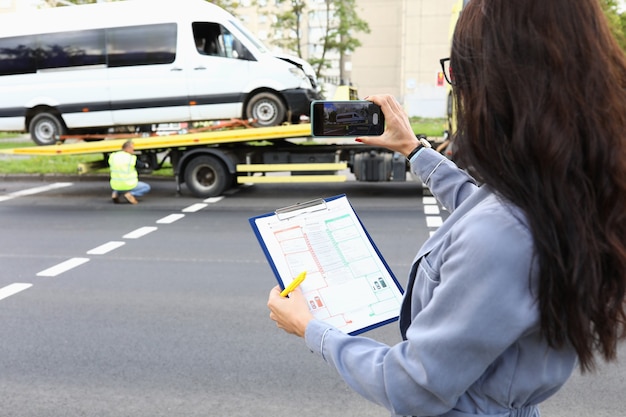 Foto mulher com documentos nas mãos tirando fotos de um carro acidentado no telefone