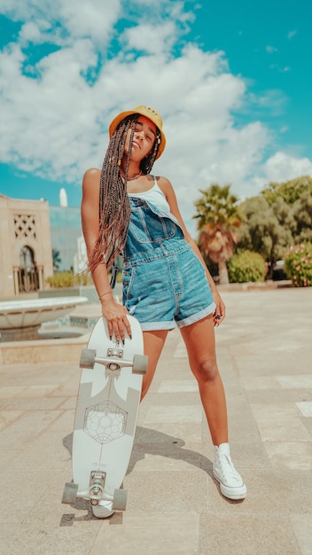 Mulher com dreadlocks em um chapéu de balde e skate posando para a câmera