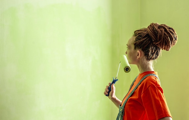 Mulher com dreadlocks em uma camiseta laranja e macacão jeans está pintando a parede com um rolo na cor verde Conceito de renovação de reparo do novo apartamento Copiar espaço